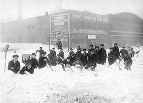 About 2 dozen young boys dressed in coats and hats all hold shovels and are standing in front of a Ford service station on the south side of Chicago during a blizzard, with snow up to their waistlines.